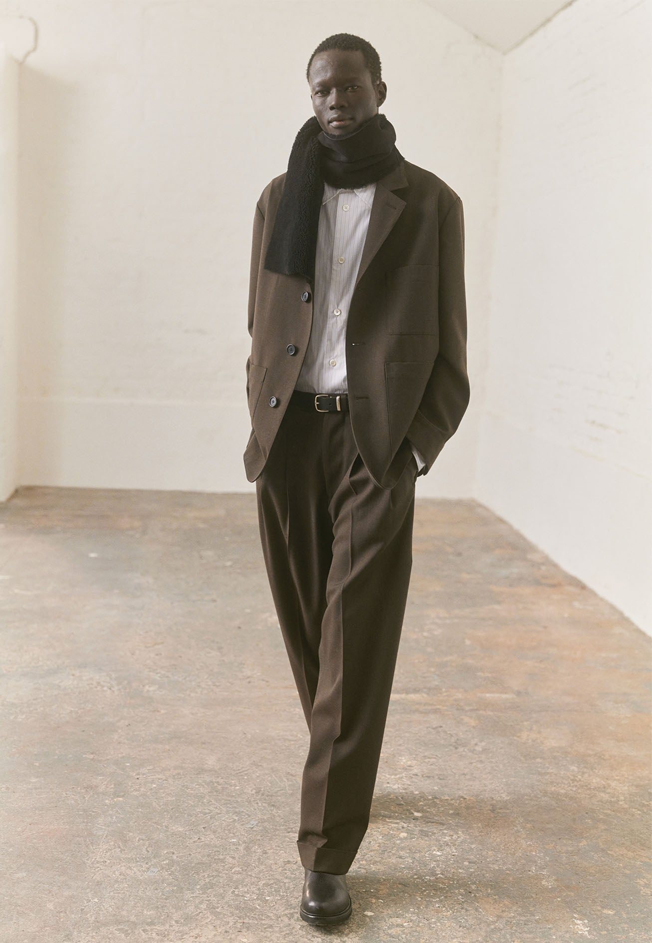 Full body image, front view of model wearing regular cut white shirt with grey stripes, chest pocket and mother of pearl buttons. Styled underneath khaki green blazer and matching trousers, with black shearling scarf and black leather boots.