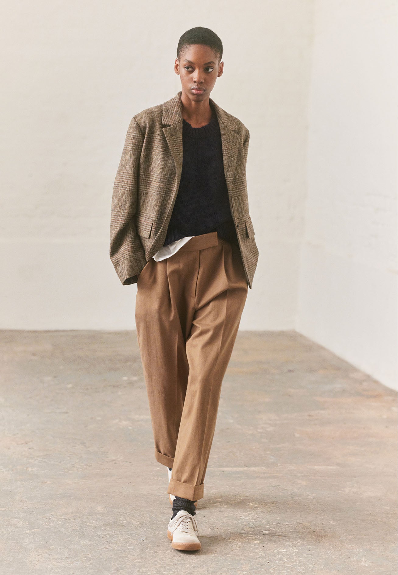 Full body image, front view of model wearing cropped green-brown glencheck blazer with horn buttons on lapel and cuffs and flap patch pockets. Styled with navy blue knitted vest, beige pleated trousers and white trainers with gum sole.