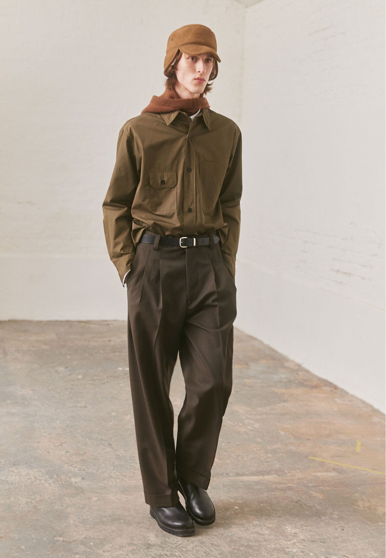 Full body image, front view of model wearing green shirt with patch pocket and button-through flap pocket on chest, raised placket with horn buttons. Styled with brown pleated trousers, black leather belt and boots and beige shearling hat.