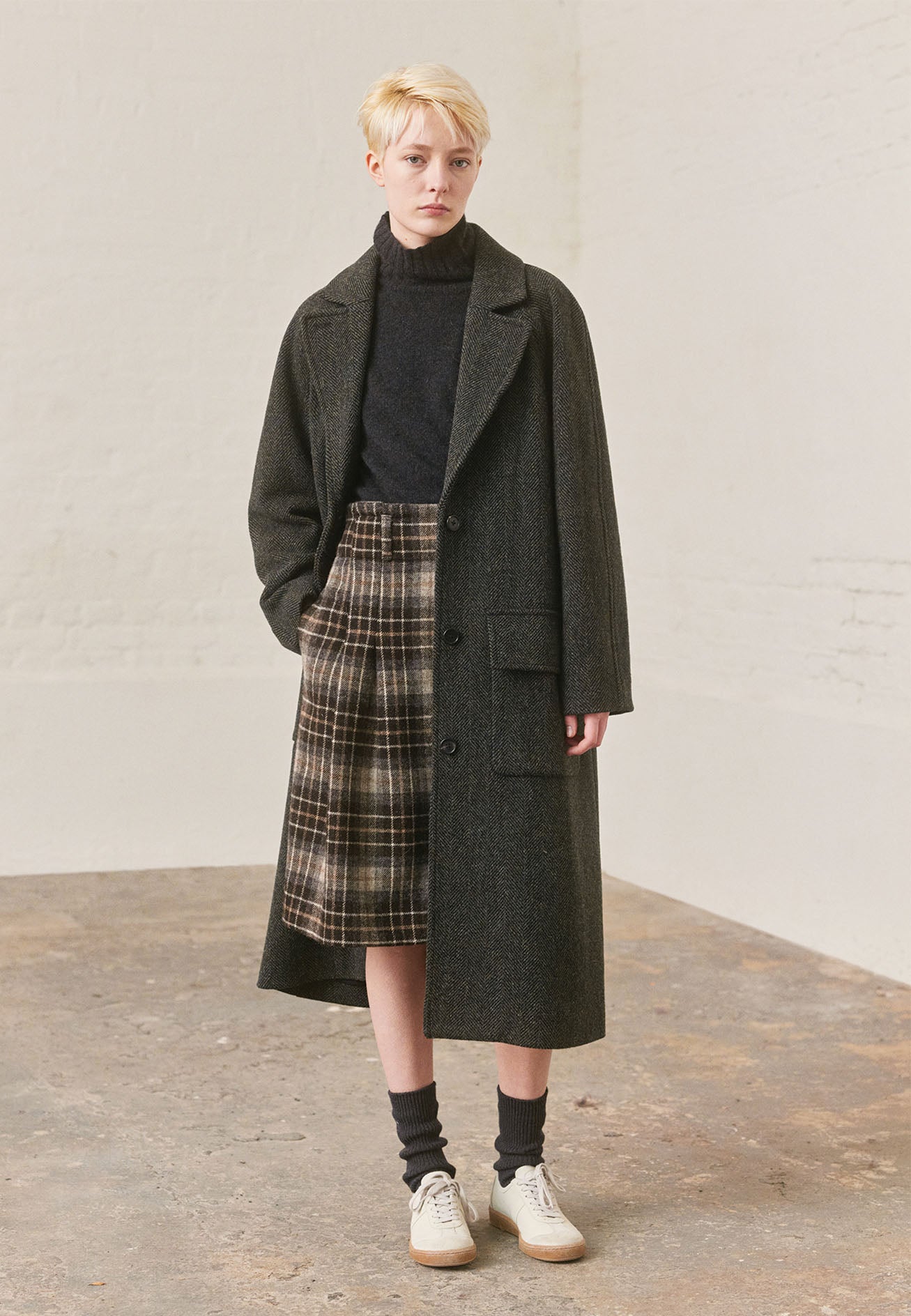 Full body image, front view of model wearing green and black herringbone wool longline overcoat with raglan sleeve, button fastening lapel and deep flap patch pockets. Styled with charcoal grey high neck jumper, brown and beige checked skirt, grey socks and white trainers with gum sole.
