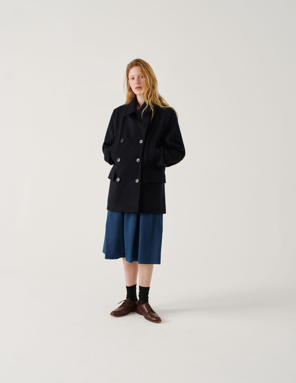 Full body image, front view of model wearing dark blue wool coat with large collar and revere, double breasted front with horn buttons, high-set single welt pockets and large flap pockets. Styled with blue midi skirt, black socks and brown leather brogues.