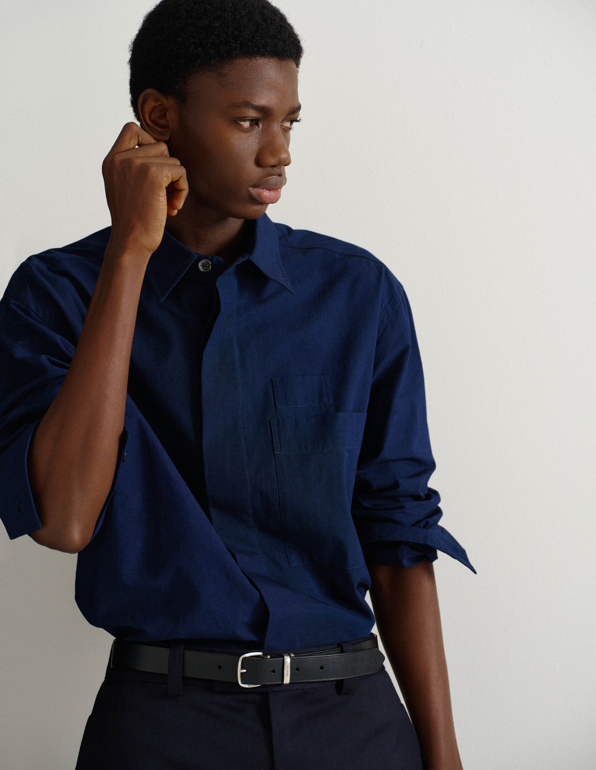 Cropped body image, front view of model wearing dark blue relaxed cotton shirt with concealed placket and mother of pearl button on collar, large patch chest pocket with internal pen pocket and vent split hem. Styled with sleeves rolled up, and with dark blue trousers and black leather belt.