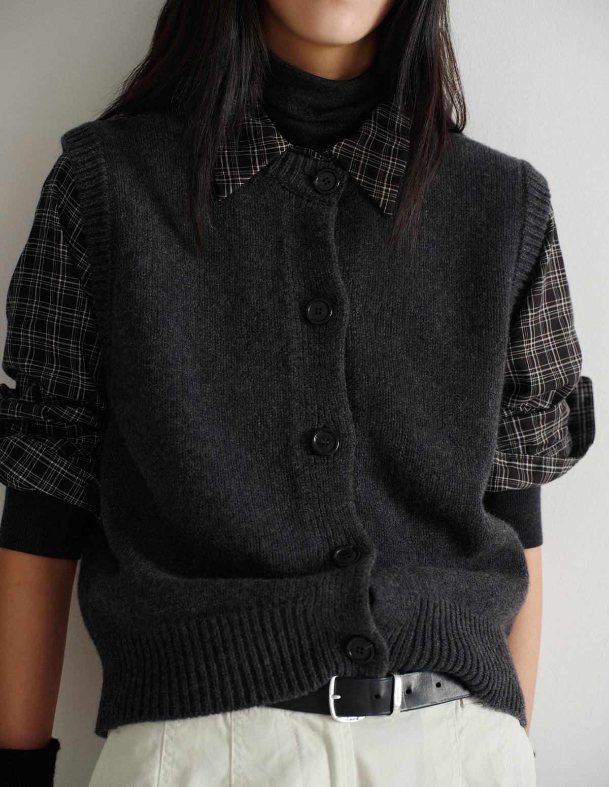 Cropped body image, front view of model wearing black and white cotton check shirt with mother of pearl buttons on placket, stepped hem with curved front and straight-cut back. Styled with sleeves rolled up under dark grey knitted gilet and over dark grey roll neck knitted top with rolled up sleeves, and with white trousers and black leather belt.