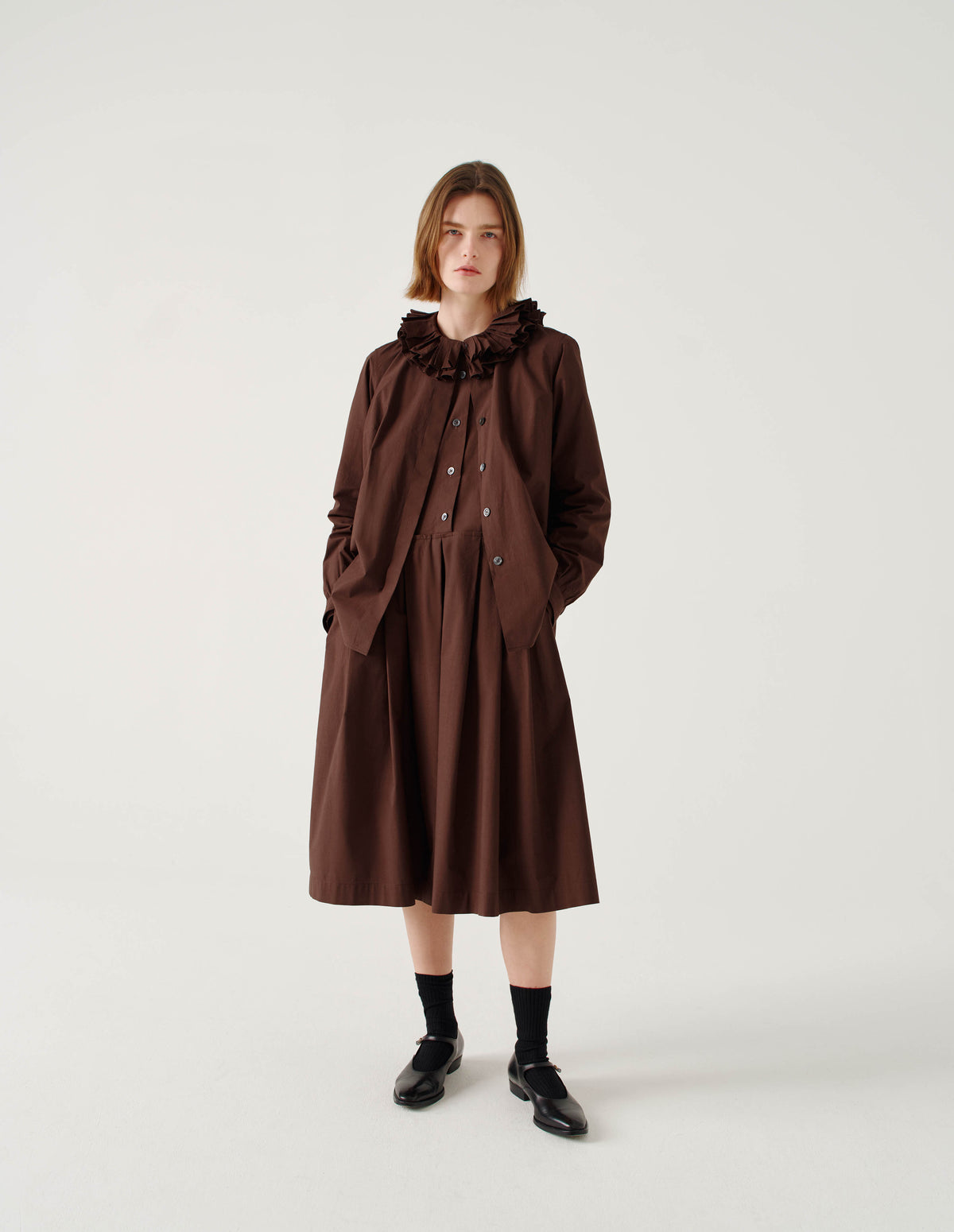 Full body image, front view of model wearing relaxed brown collarless cotton midi dress with mother of pearl buttons on half placket, long sleeves with shirt cuffs, and pleated skirt with concealed side pockets. Styled under matching  ruffle shirt, with shirt buttons undone, and with black socks and black leather shoes.