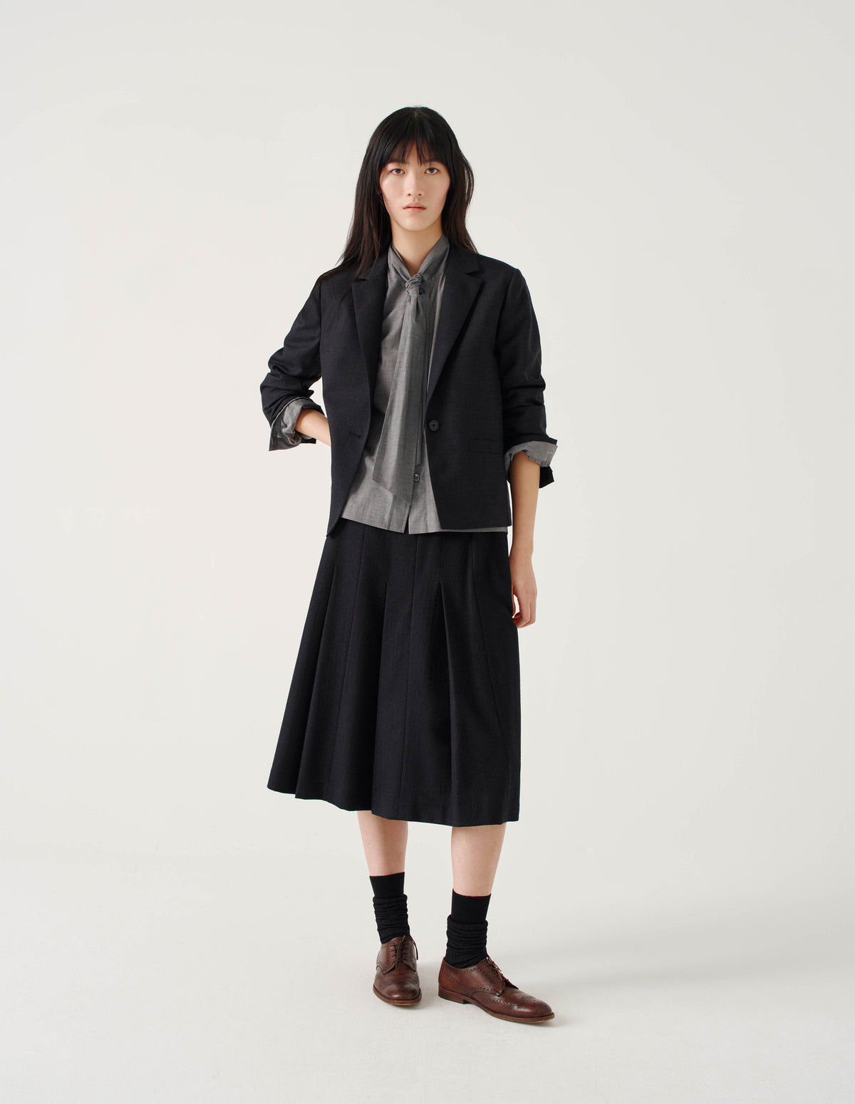 Full body image, front view of model wearing below knee length black wool skirt with dark grey stripes, pleats, narrow horn button-through waistband and side zip fly. Styled with grey front tie shirt under dark grey blazer with sleeves rolled up, and with black socks and brown leather brogues.