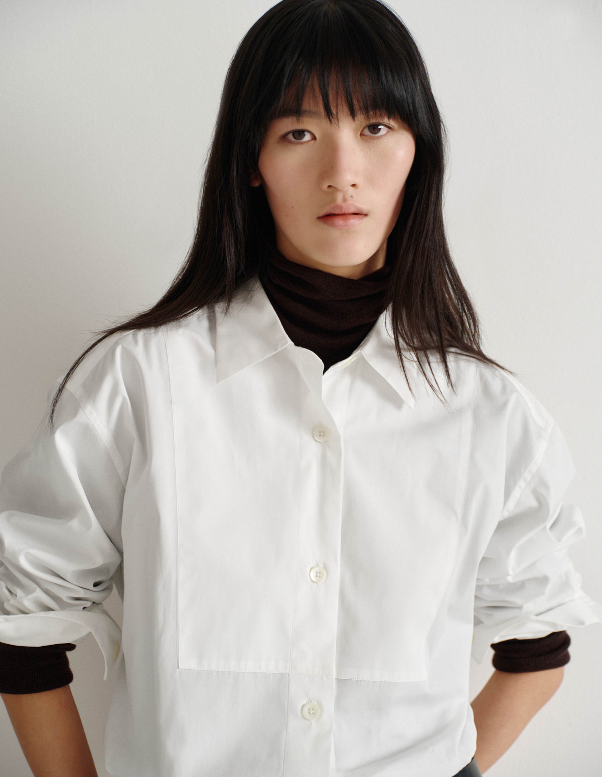Cropped body image, front view of model wearing dark brown merino knitted top with fully fashioned roll neck and armhole, and with ribbed neck, sleeves, cuff and hem. Styled under white bib front shirt, sleeves rolled up.