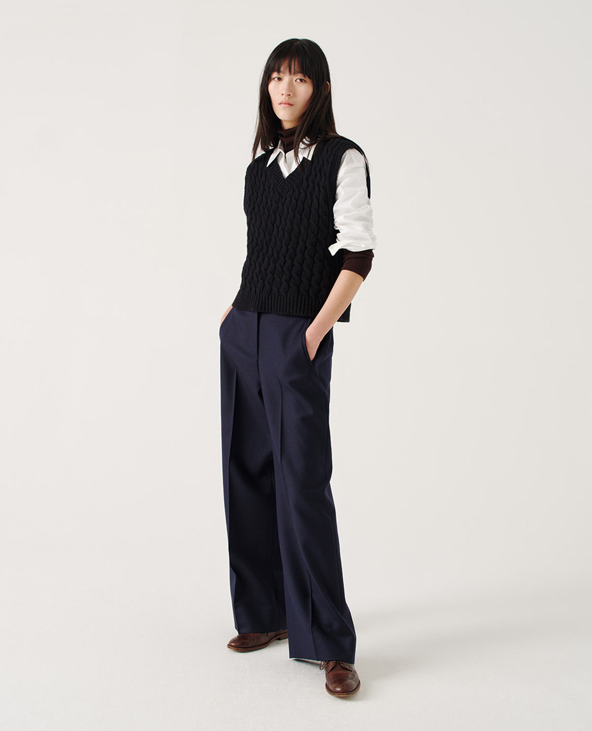 Full body image, front view of model wearing dark blue high waisted wool trousers with wide leg, belt loops on wide waistband, zip fly and horn button fastening, side adjusters and slanted front welt pockets. Styled with black slipover over dark brown roll neck and white shirt, with sleeves rolled up, and brown leather brogues.
