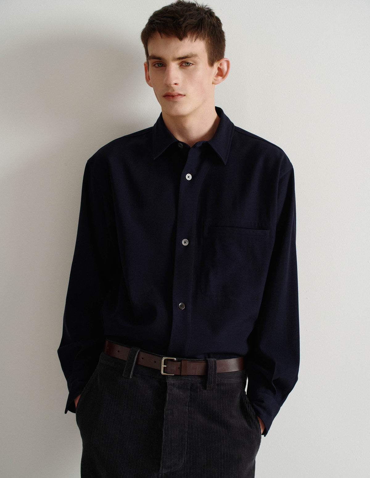 Cropped image, front view of model wearing relaxed dark blue wool shirt with mother of pearl buttons and welt chest pocket. Styled with dark grey corduroy trousers and brown leather belt.