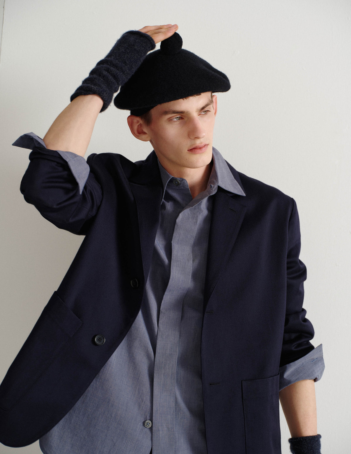 Cropped body image, front view of male model wearing dark blue merino cashmere fingerless mittens with contrasting blue and grey flecks. Styled with black beret and dark blue blazer over blue shirt with sleeves rolled up.