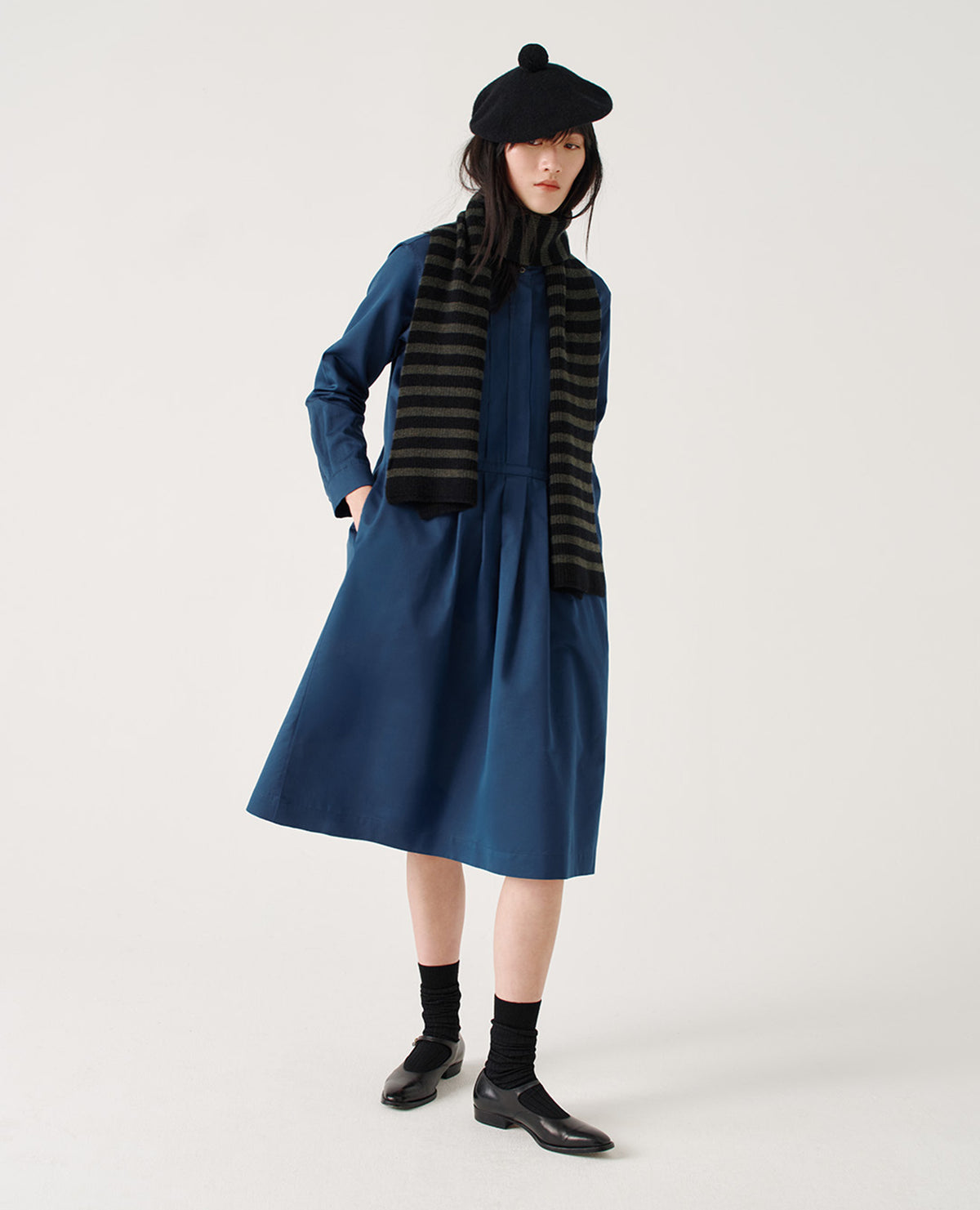 Full body image, front view of model wearing blue cotton silk collarless midi dress with concealed half placket on shirt front with single mother of pearl button fastening, shirt sleeves with mother of pearl buttons on narrow cuffs, side seam pockets, deep stitch-through pleats on front bib and skirt, and split hems. Styled with black tam o'shanter, black and grey striped scarf, black socks and black leather shoes.