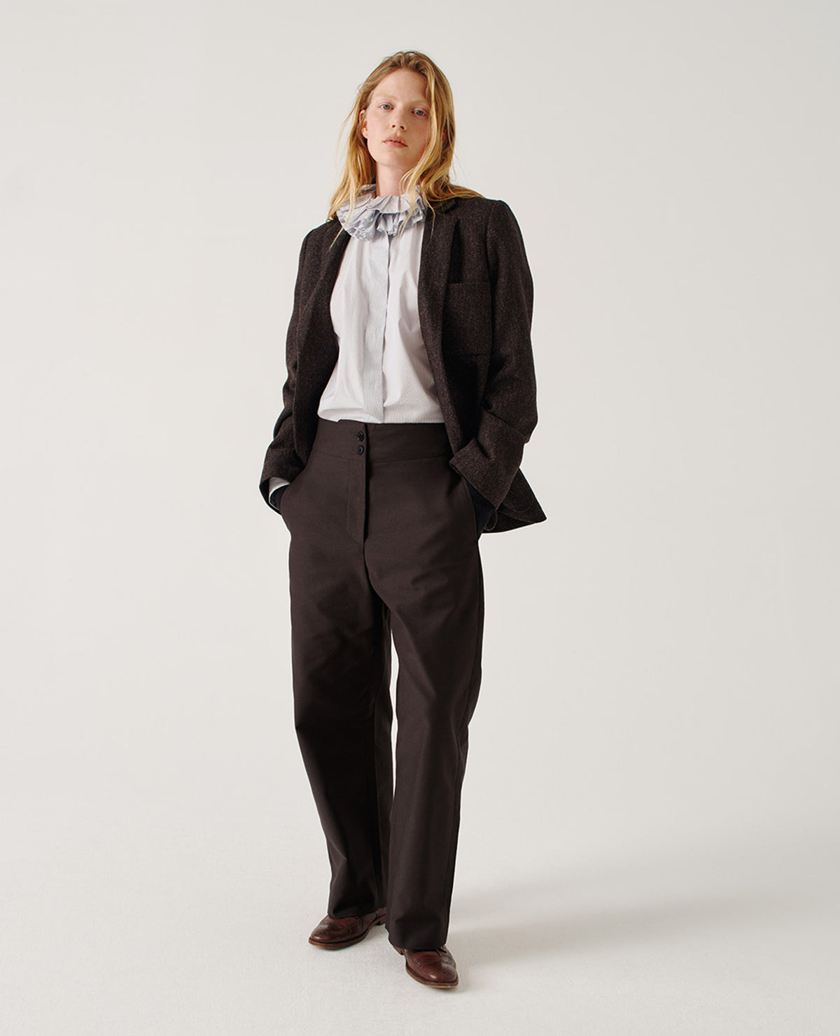 Full body image, front view of model wearing brown and black herringbone wool blazer with narrow collar and horn button-through revere, three bluff patch pockets, rounded details on hem and pockets. Styled over light grey frilled shirt, tucked in, and with dark grey trousers and brown leather brogues.