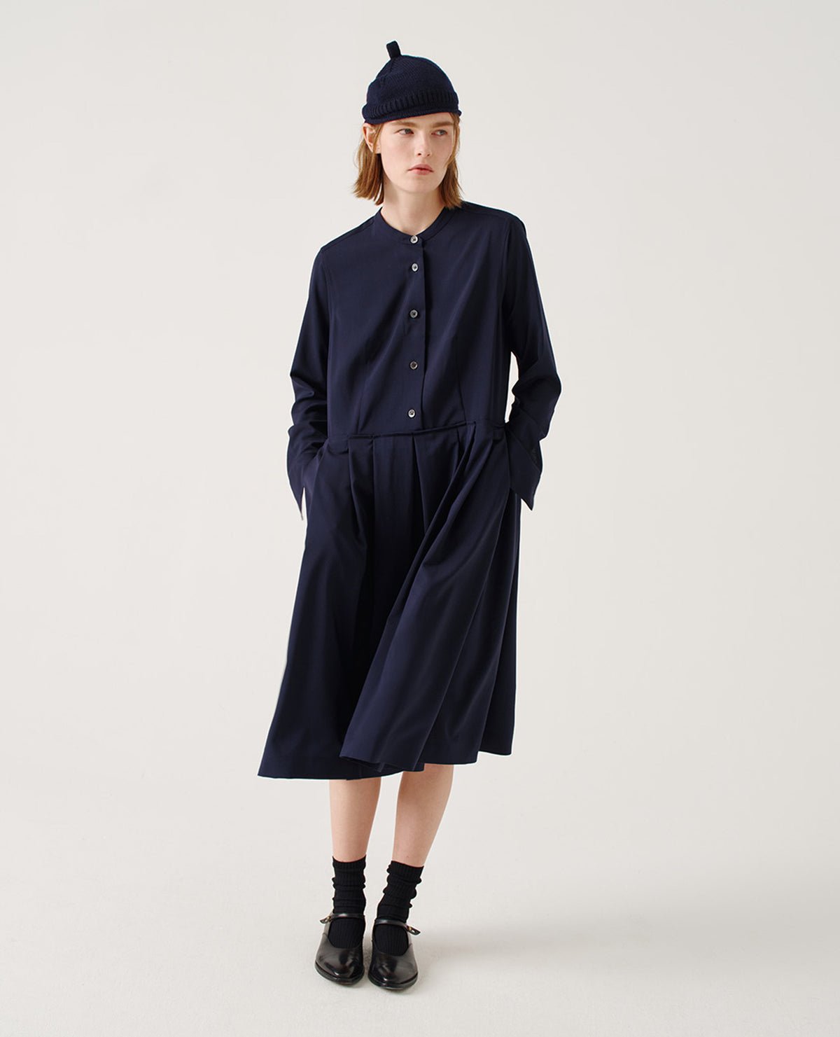 Full body image, front view of model wearing relaxed dark blue collarless wool midi dress with mother of pearl buttons on half placket, long sleeves with shirt cuffs, and pleated skirt with concealed side pockets. Styled with dark blue beanie, black socks and black leather shoes.