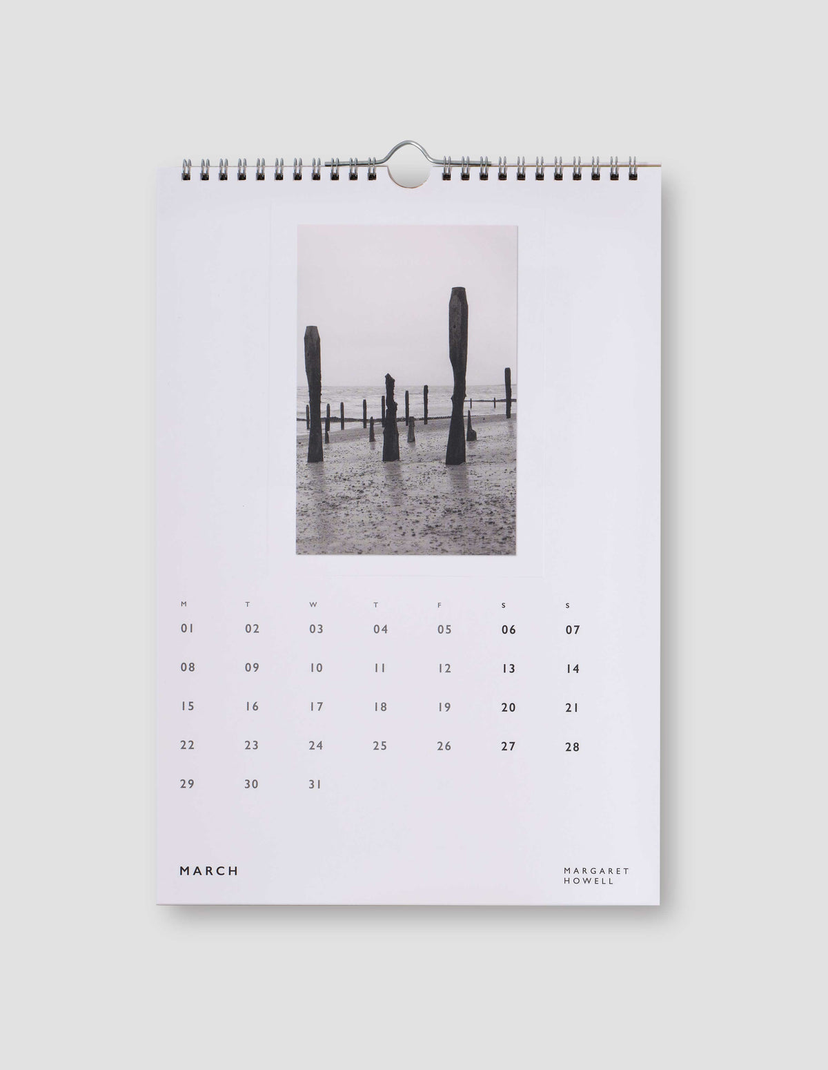 An image of a white page from the calendar featuring a black and white photograph of groynes on a beach, with days of the month under the image. Flat lay image on a light grey background.