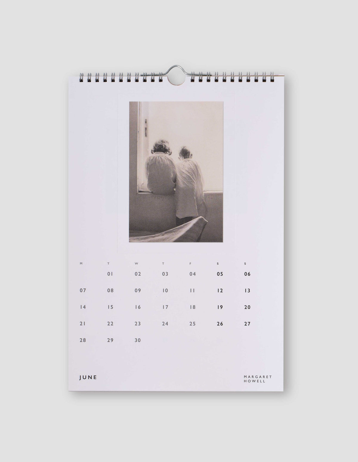 An image of a white page from the calendar featuring a black and white photograph of two children with their backs to the camera, and with days of the month under the image. Flat lay image on a light grey background.