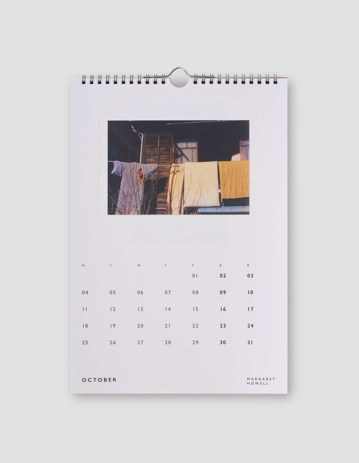 An image of a white page from the calendar featuring a  colour photograph blankets and clothes hanging in front of a house, and with days of the month under the image. Flat lay image on a light grey background.