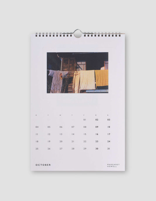 An image of a white page from the calendar featuring a  colour photograph blankets and clothes hanging in front of a house, and with days of the month under the image. Flat lay image on a light grey background.