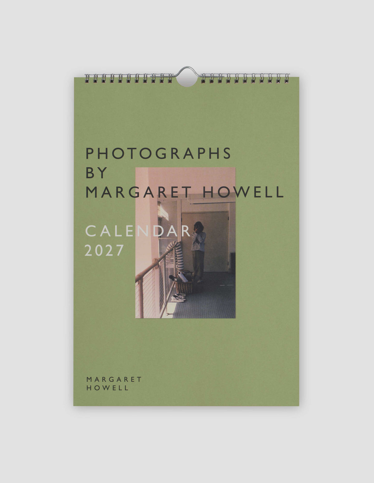 Green front cover of the annual calendar titled Photographs by Margaret Howell, Calendar 2027, over a black and white photograph of a woman holding a camera, and with Margaret Howell logo in the left corner. Flat lay image on a light grey background.