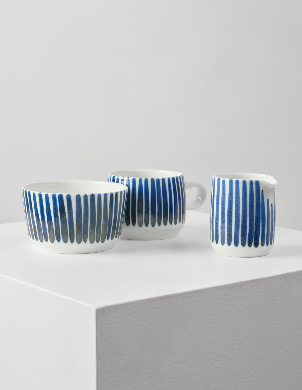 Front view of white porcelain teacup, pouring jug and bowl, all with hand painted vertical blue stripes.