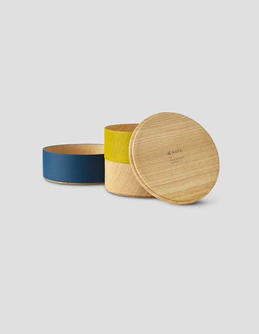 Front flat lay of a round wooden container with a light natural wood lid resting against yellow and natural wood tiers, with 'HATA for Margaret Howell' printed on the inside of the lid. The dark blue tier is sat to the left side.