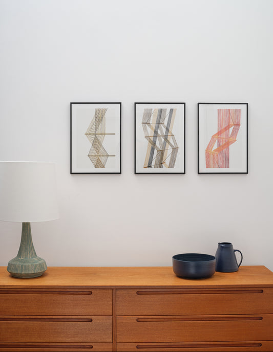 Three framed colourful prints of a Peter Collingwood macrogauze woven wall hangings next to each other on a pale wall hanging above a wooden sideboard with lamp and ceramics.
