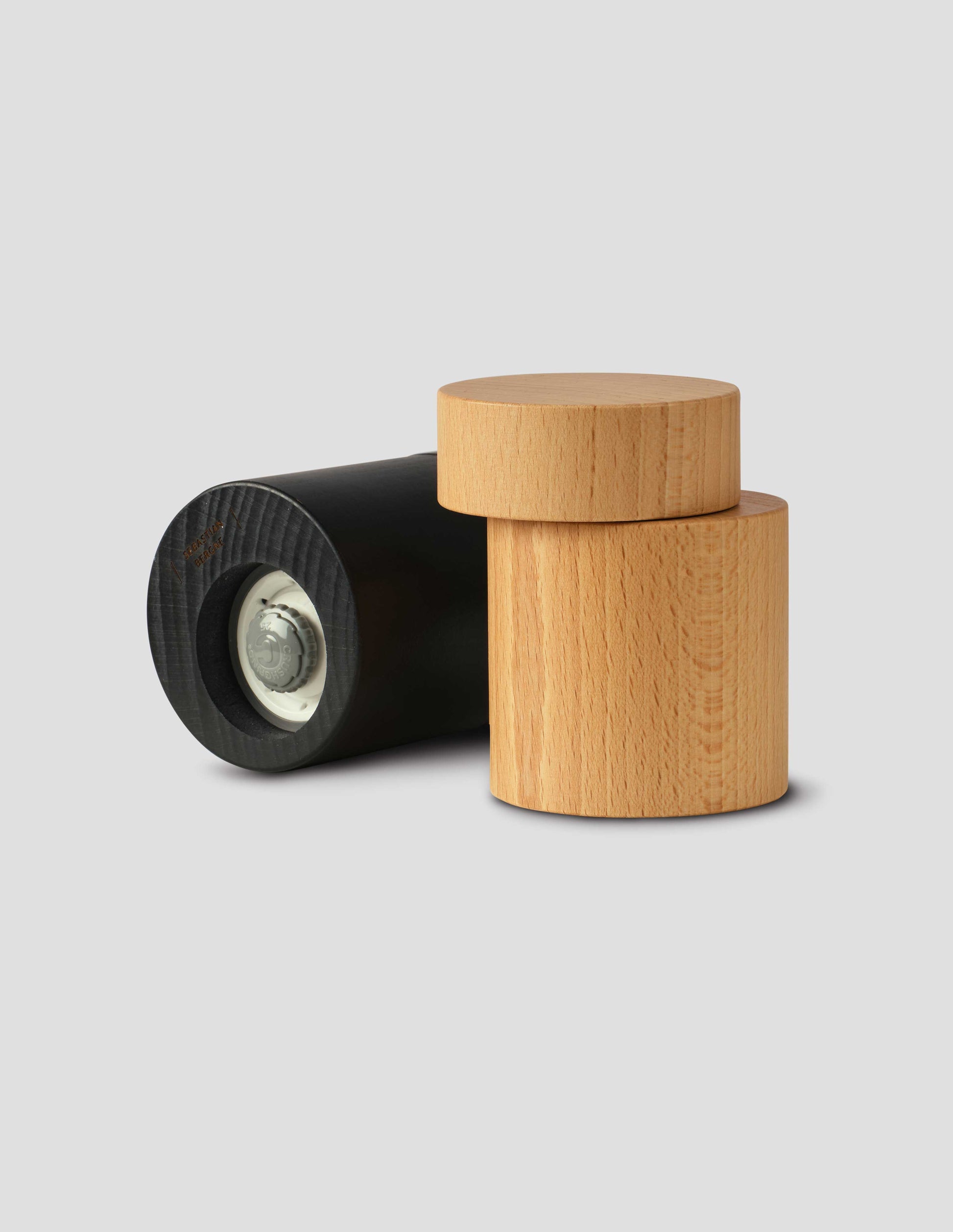 A group shot of one matte black cylindrical salt and pepper mill lay on it's side, next to one wooden cylindrical salt and pepper mill showing the grinding functionality.