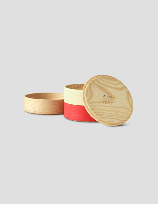 Front flat lay of a round wooden container with a light natural wood lid resting against cream and red tiers, with The 'HATA for Margaret Howell' printed on the inside of the lid. The natural wood tier is sat to the left side.