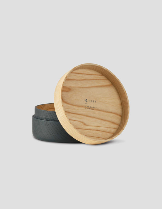 Front flat lay of a round wooden container with a light natural wood lid resting against the matte black wooden base. With The 'HATA for Margaret Howell' printed on the inside of the lid.