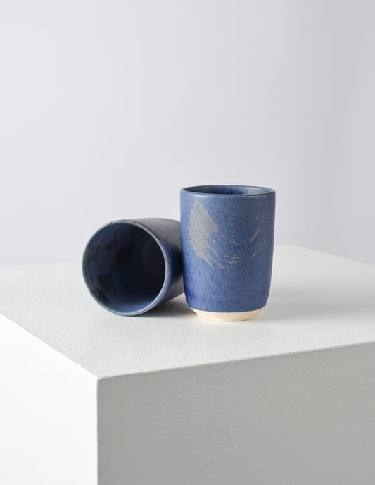 A pair of tall indigo beakers, one on its side so you can see the variation of the blue inner glaze with dark flashes.