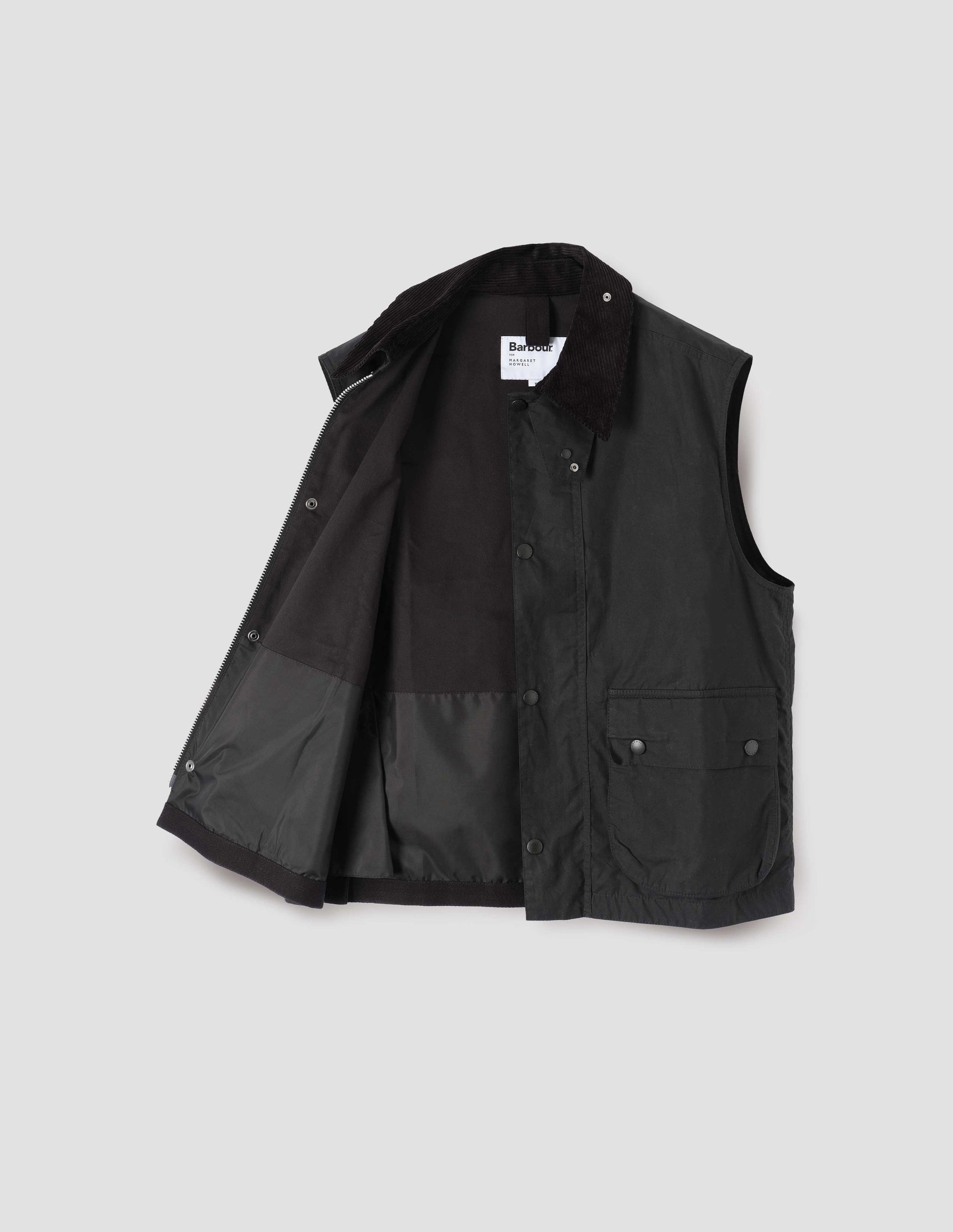 Black cotton drill lined gilet with storm tab on corduroy collar, two-way zip fastening and press stud fastening storm flap, large bellows pockets with press stud fastening envelope closure. Open flat lay view.