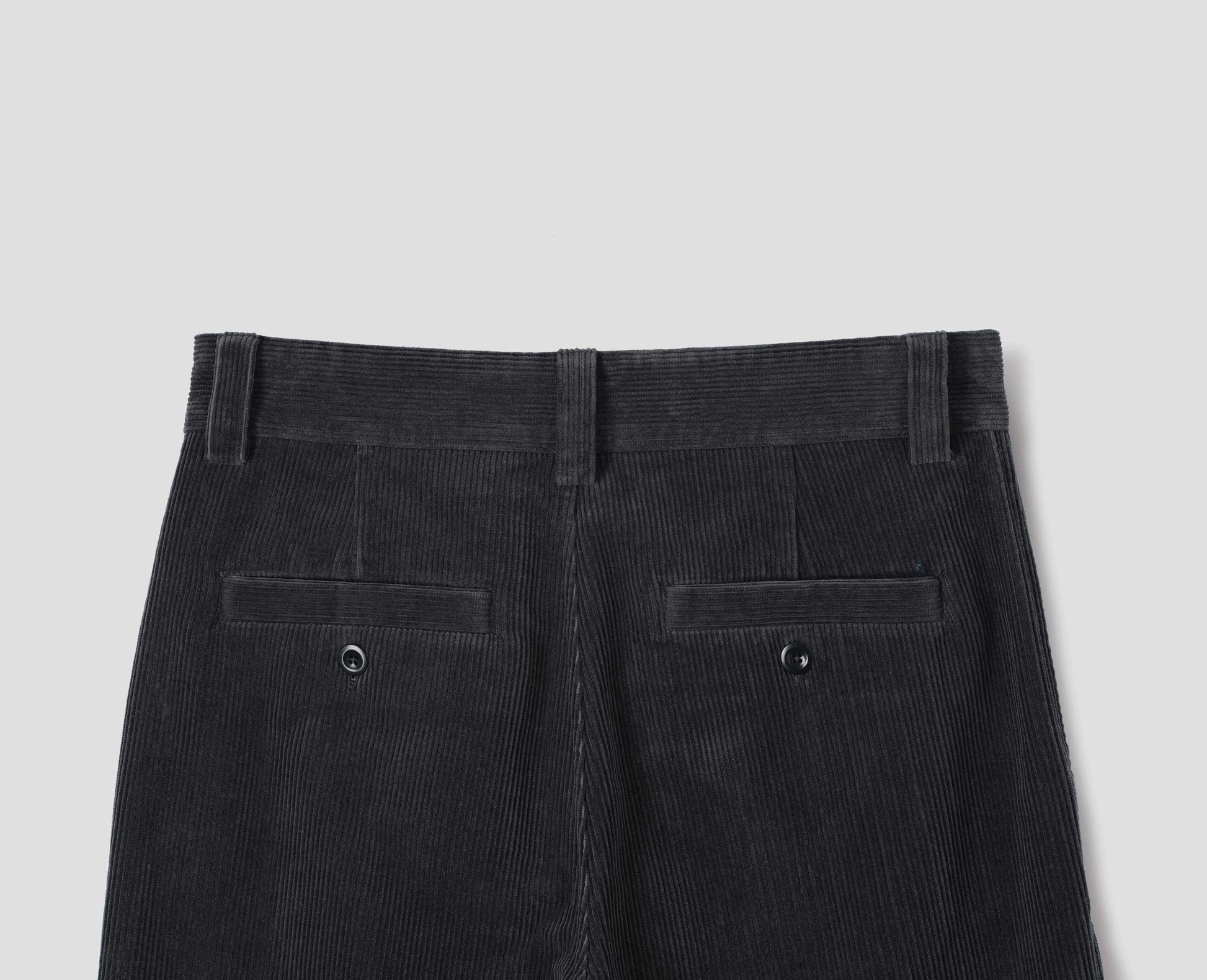 Dark grey cotton corduroy trousers with belt loops and two welt back button fastening pockets. Back flat lay view.