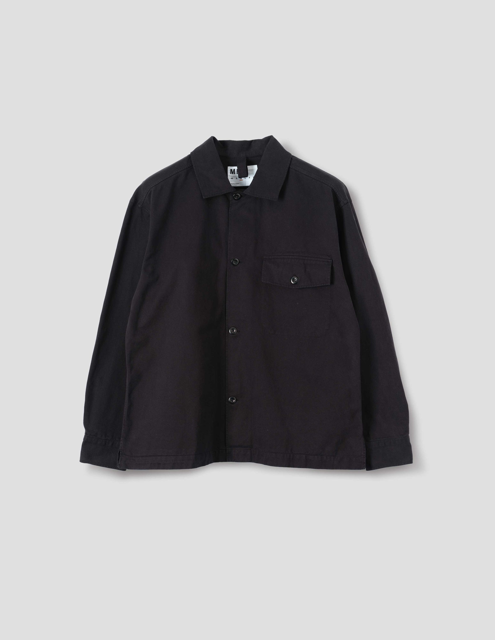 Black cotton overshirt, with a grown-on, button-fastening placket, spread collar, and welt chest pockets with flaps and button closure, finished with shirt cuffs. Front flat lay view. 