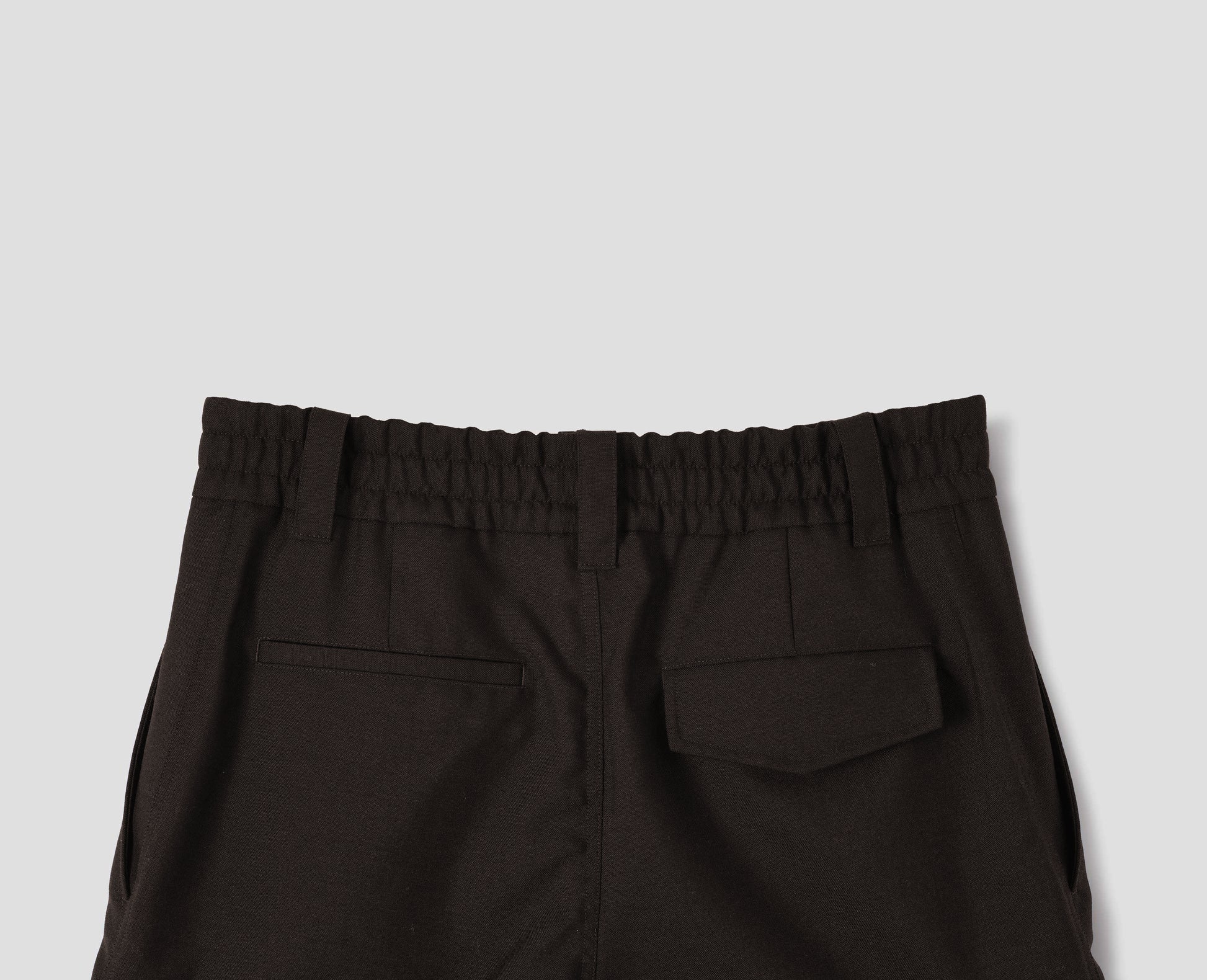 Dark brown wool trousers with half-elasticated waist with belt loops, slant side pockets, and one welt back pocket and one flap back pocket with concealed button fastening. Back flat lay view.