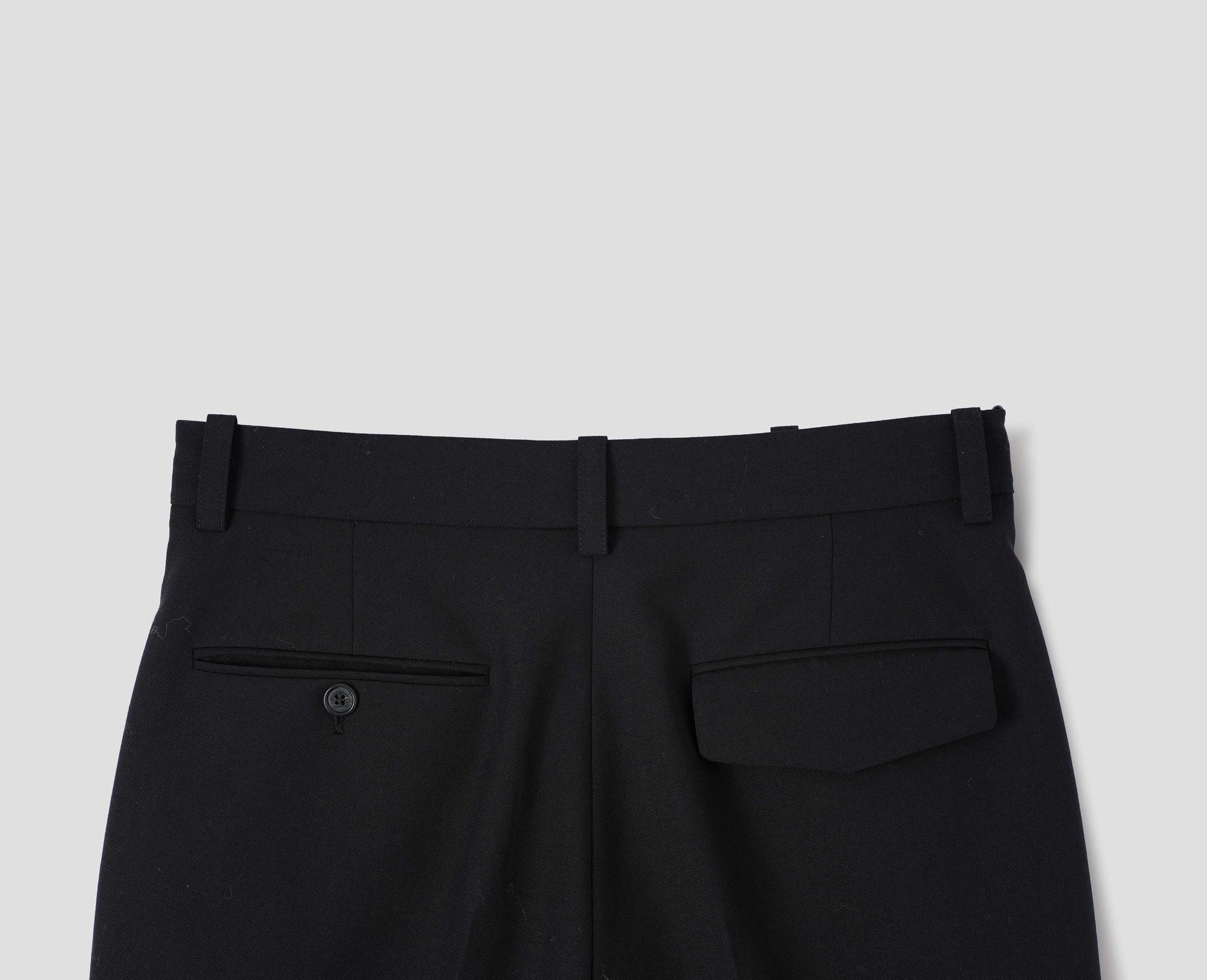 Black wool trousers with belt loops, one jetted flap back pocket and one horn button through jetted back pocket. Back flat way view.