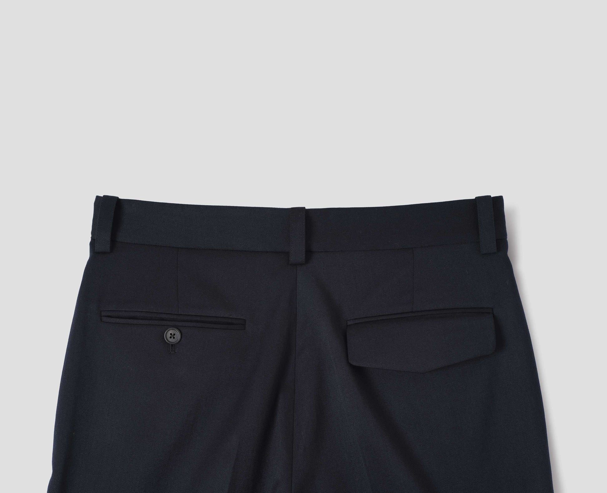 Dark blue wool cotton trousers with belt loops, one jetted flap back pocket and one horn button through jetted back pocket. Back flat way view.
