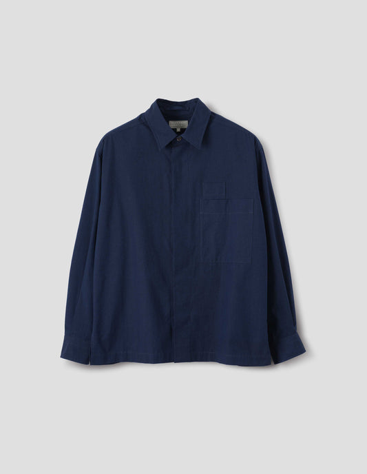 Dark blue cotton shirt with concealed placket and mother of pearl button on collar, large patch chest pocket with internal pen pocket and vent split hem. Front flat lay view.
