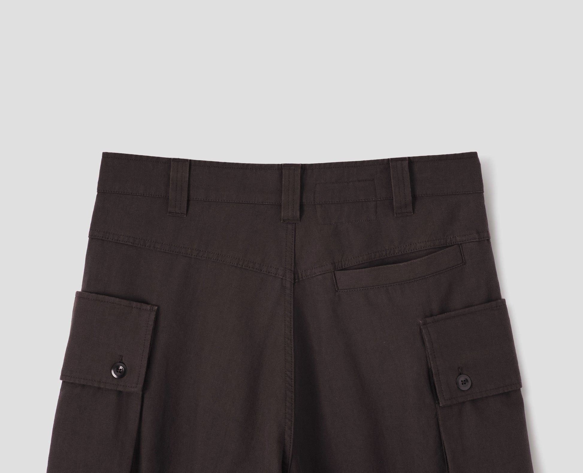 Dark brown cotton drill cargo trousers with deep flap button fastening patch pocket, belt loops and welt pocket on back yoke. Back flat lay view.