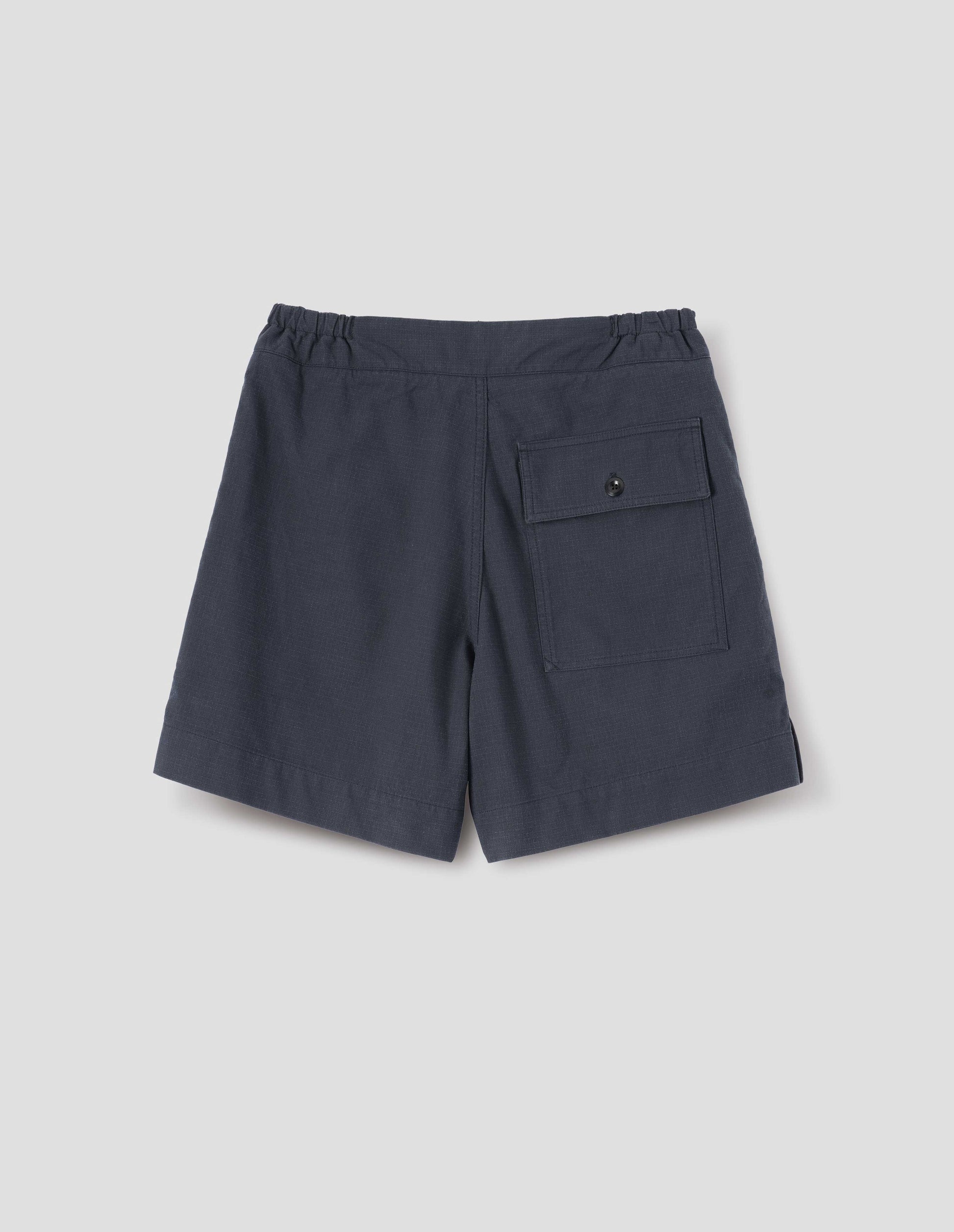 Dark blue shorts with side-elasticated waistband, corozo brace button-through back flap patch pocket and side split hem. Back flat lay view.