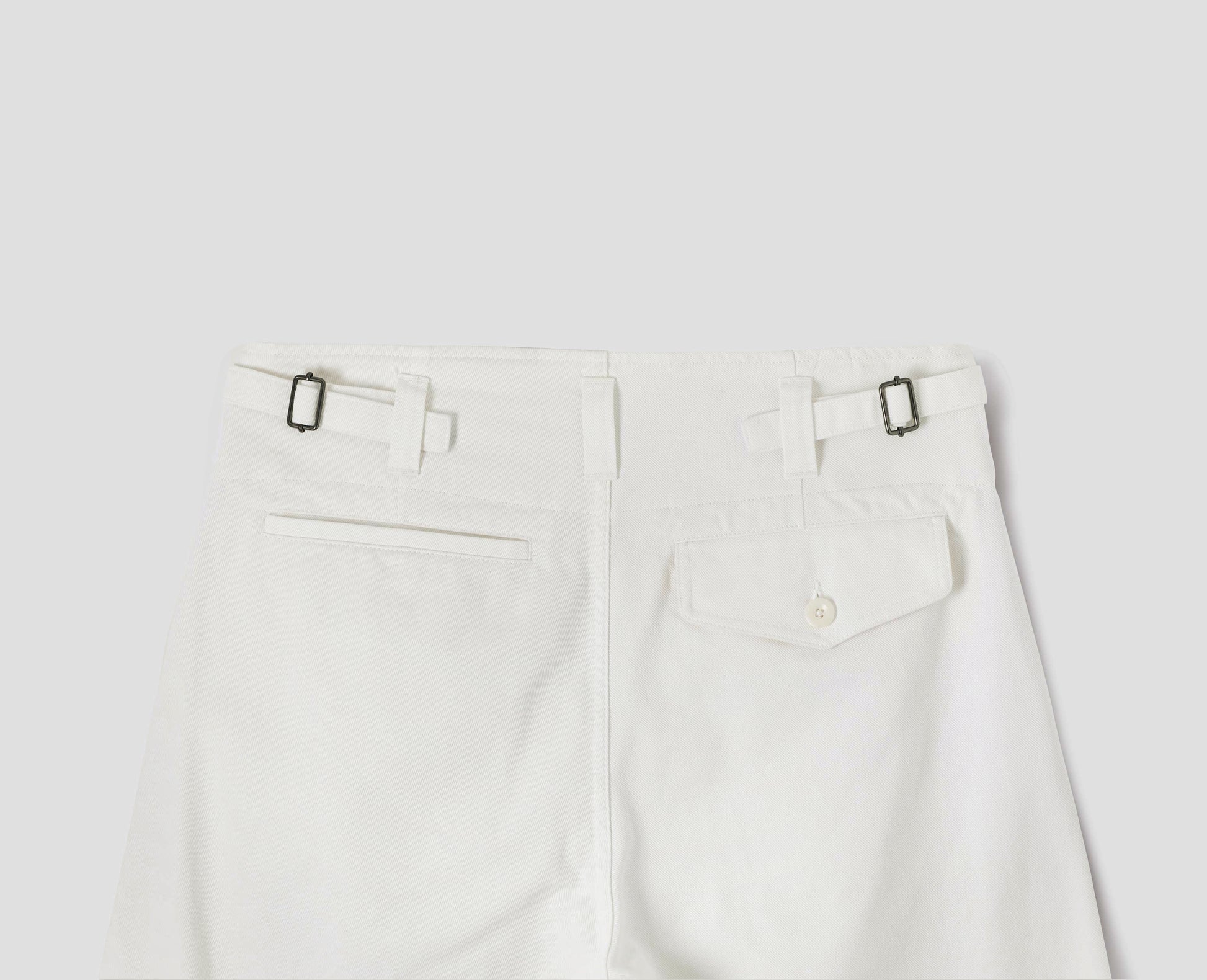 White cotton drill trousers with belt loops, side adjustors, welt back and flap back pockets. Back flat lay view.