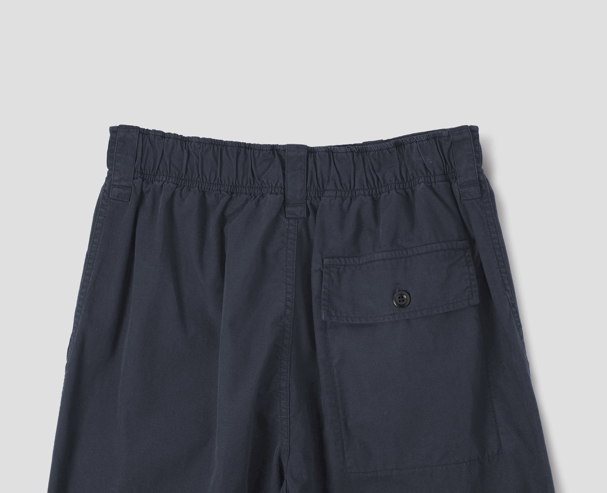 Dark blue trousers with sewn-in patch pockets, half-elasticated waistband, belt loops and corozo brace button fastening flap back pocket. Back flat lay view. 