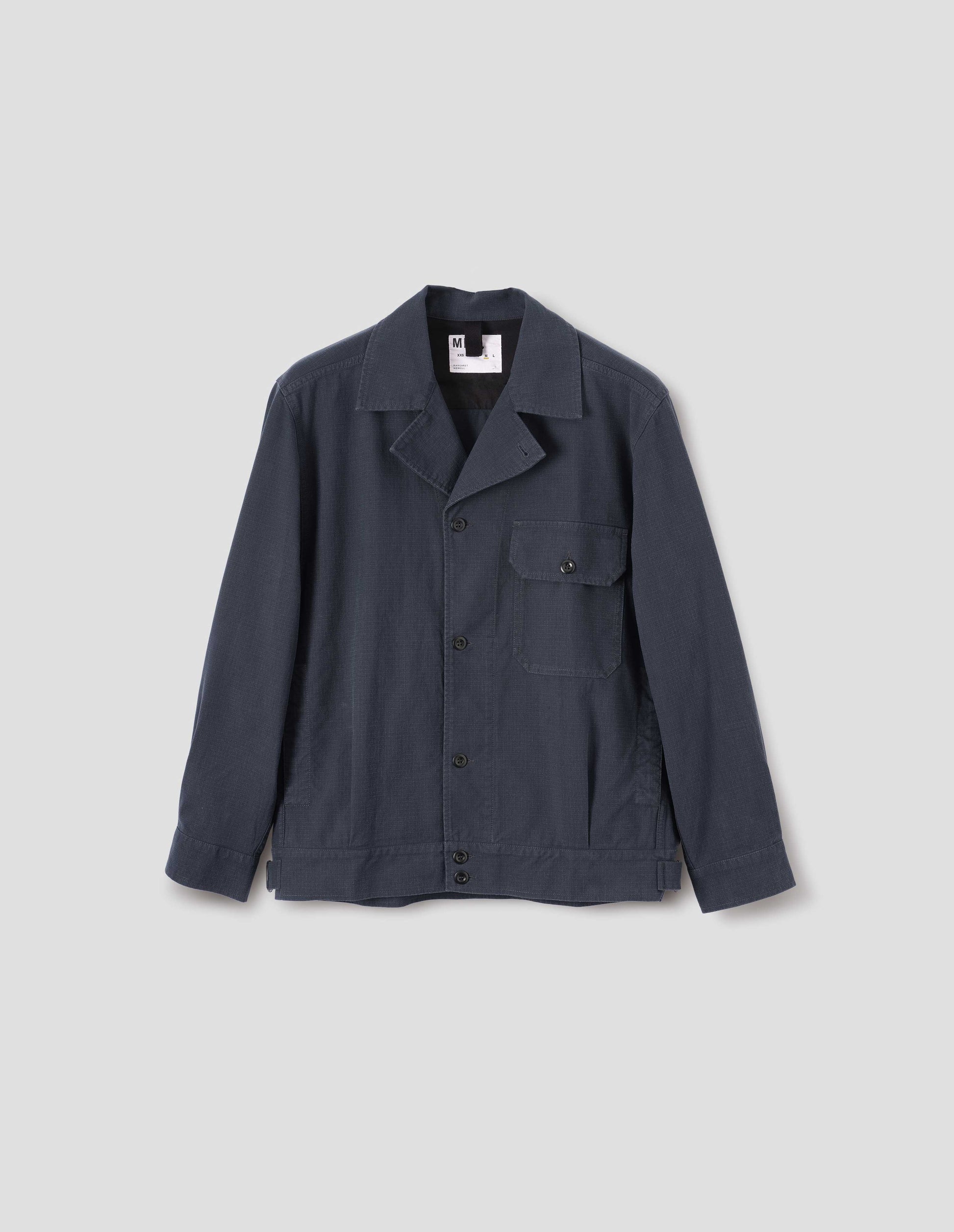 Dark blue jacket with corozo brace button fastening lapel, chest flap pocket, concealed side entry pockets and shirt cuffs, small pleats at hem and side adjustors. Front flat lay view.