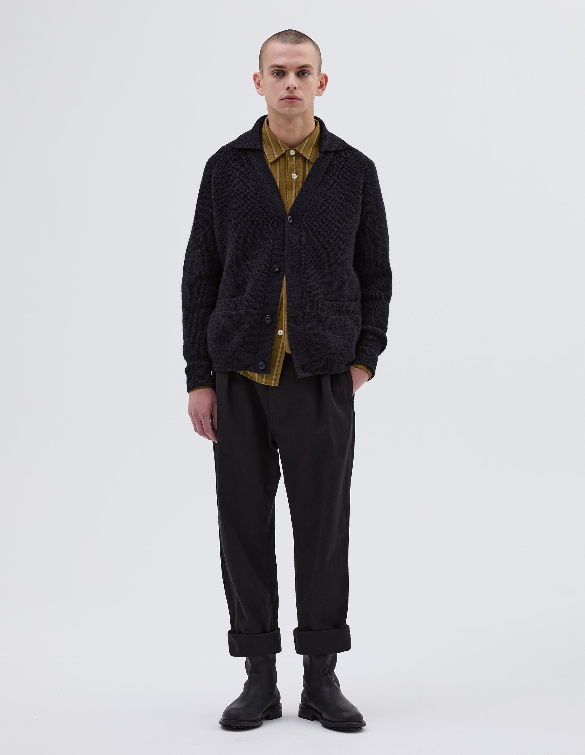 Full body image, front view of model wearing dark navy blue collared cardigan, with contrast rib on collar, placket, cuff and hem. Finished with corozo button closure and patch pockets. Styled with yellow striped shirt, black trousers and black leather boots.