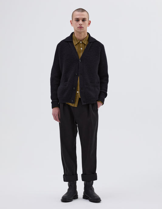 Full body image, front view of model wearing dark navy blue collared cardigan, with contrast rib on collar, placket, cuff and hem. Finished with corozo button closure and patch pockets. Styled with yellow striped shirt, black trousers and black leather boots.