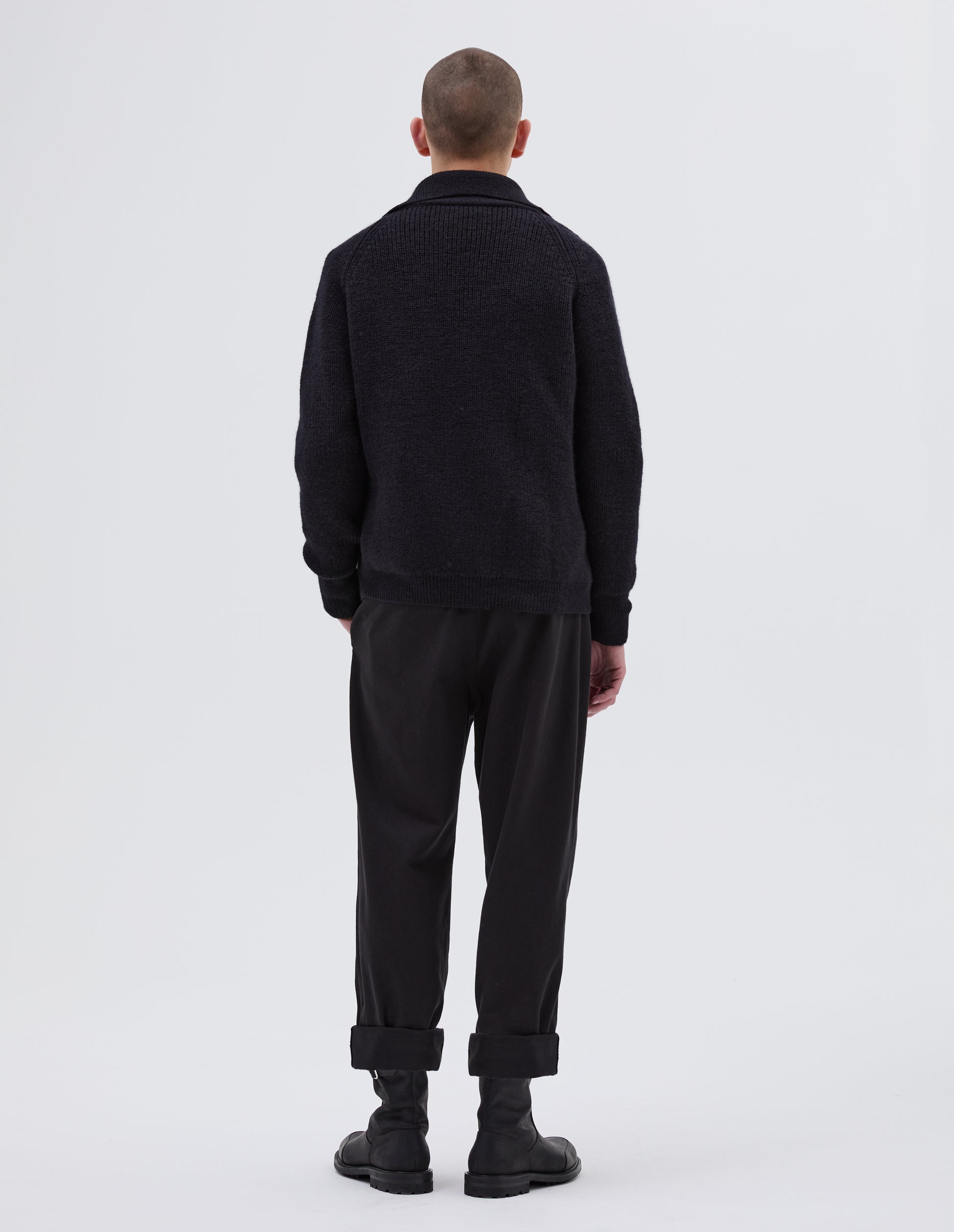Full body image, back view of model wearing dark navy blue collared cardigan, with contrast rib on collar, placket, cuff and hem. Styled with black trousers and black leather boots.