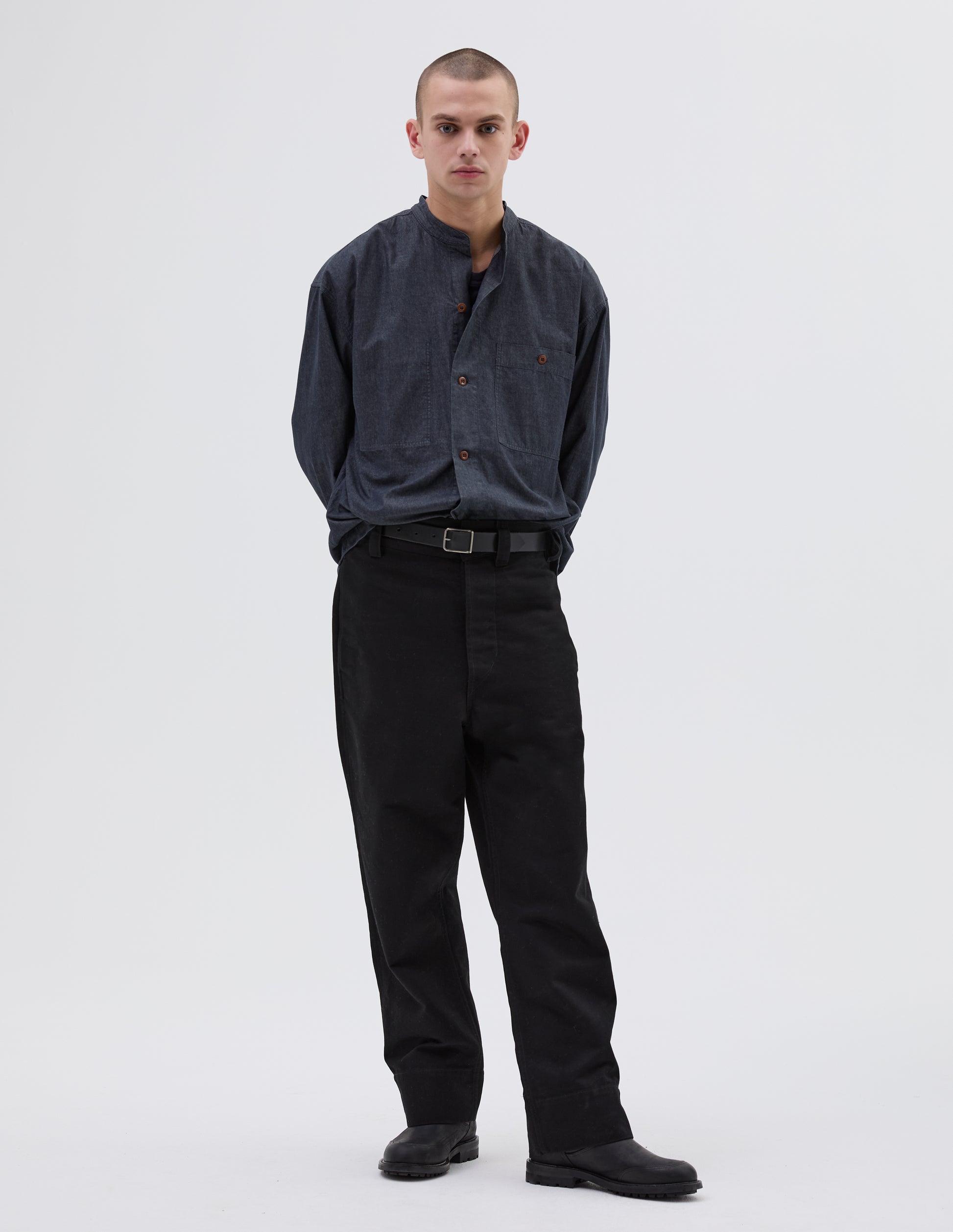 Full body image, front view of model wearing black cotton trousers, belt loops and slant pockets. Styled with indigo blue shirt, black leather belt and black leather boots.