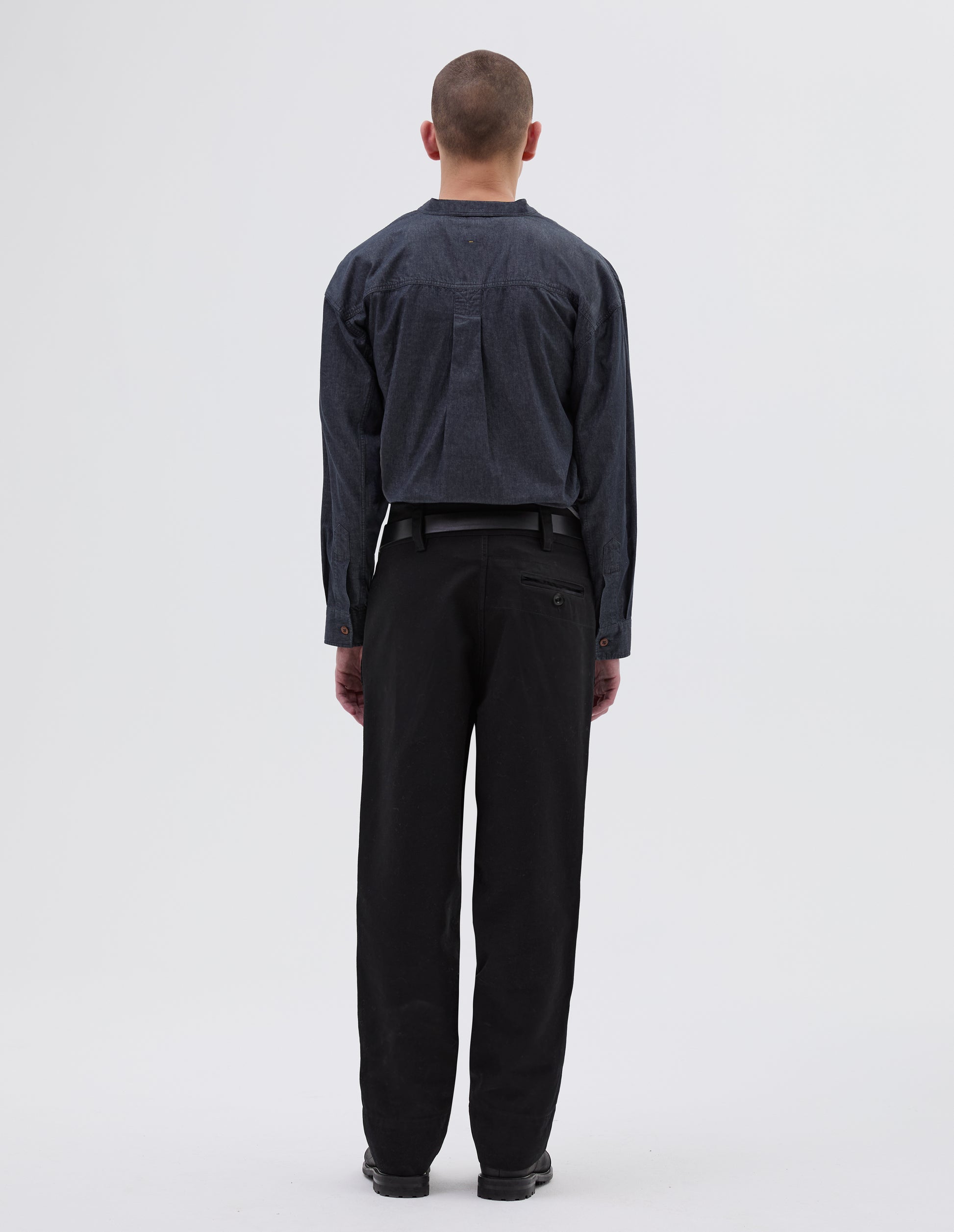 Full body image, back view of model wearing black cotton trousers, with belt loops, button through jet pocket with stitch reinforcement detailing. Styled with indigo blue shirt, black leather belt and black leather boots.