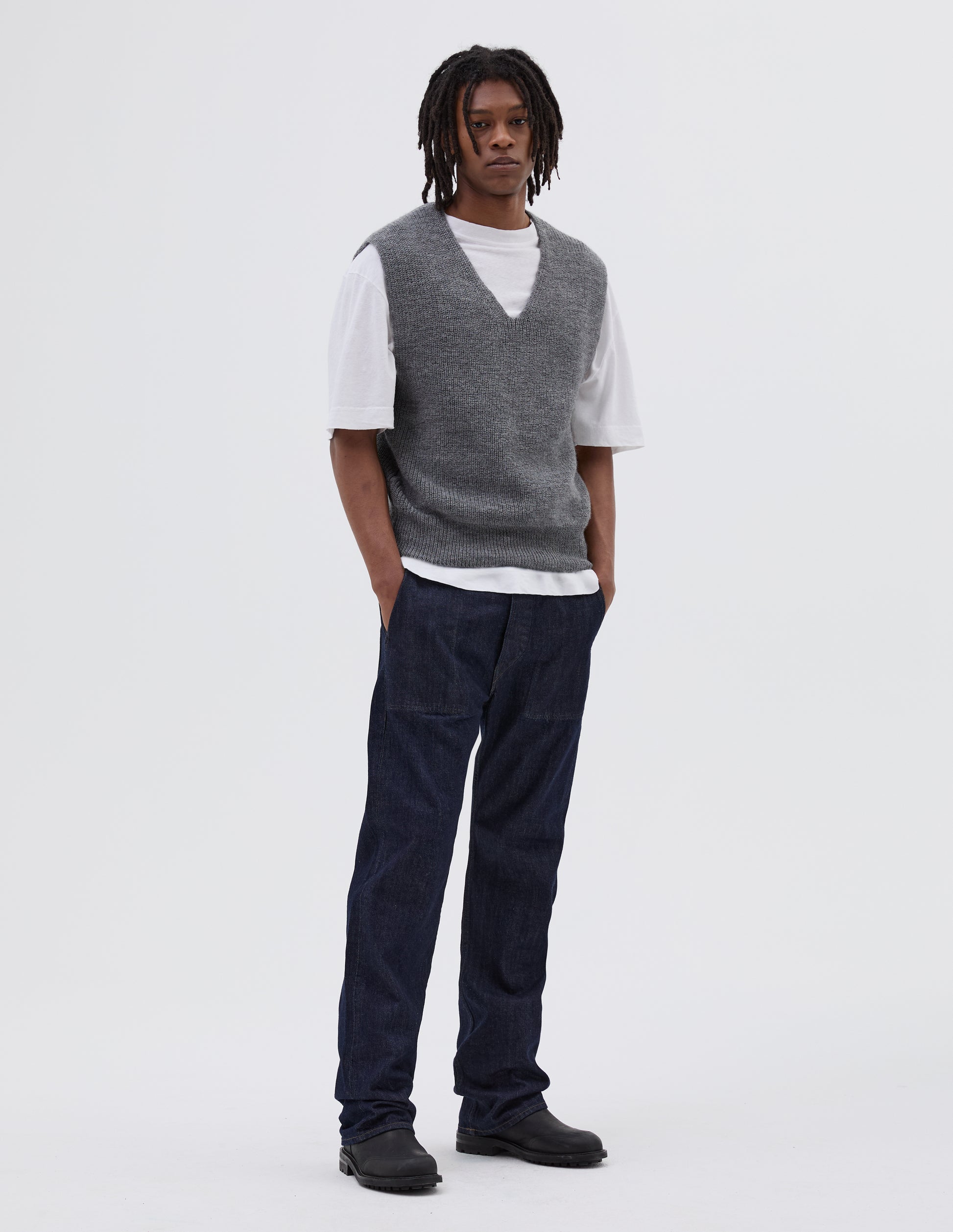 Full body image, front view of model wearing grey sleeveless ribbed v neck slipover jumper, with contrast rib on hem. Styled over white t shirt, with indigo denim jeans and black leather boots.