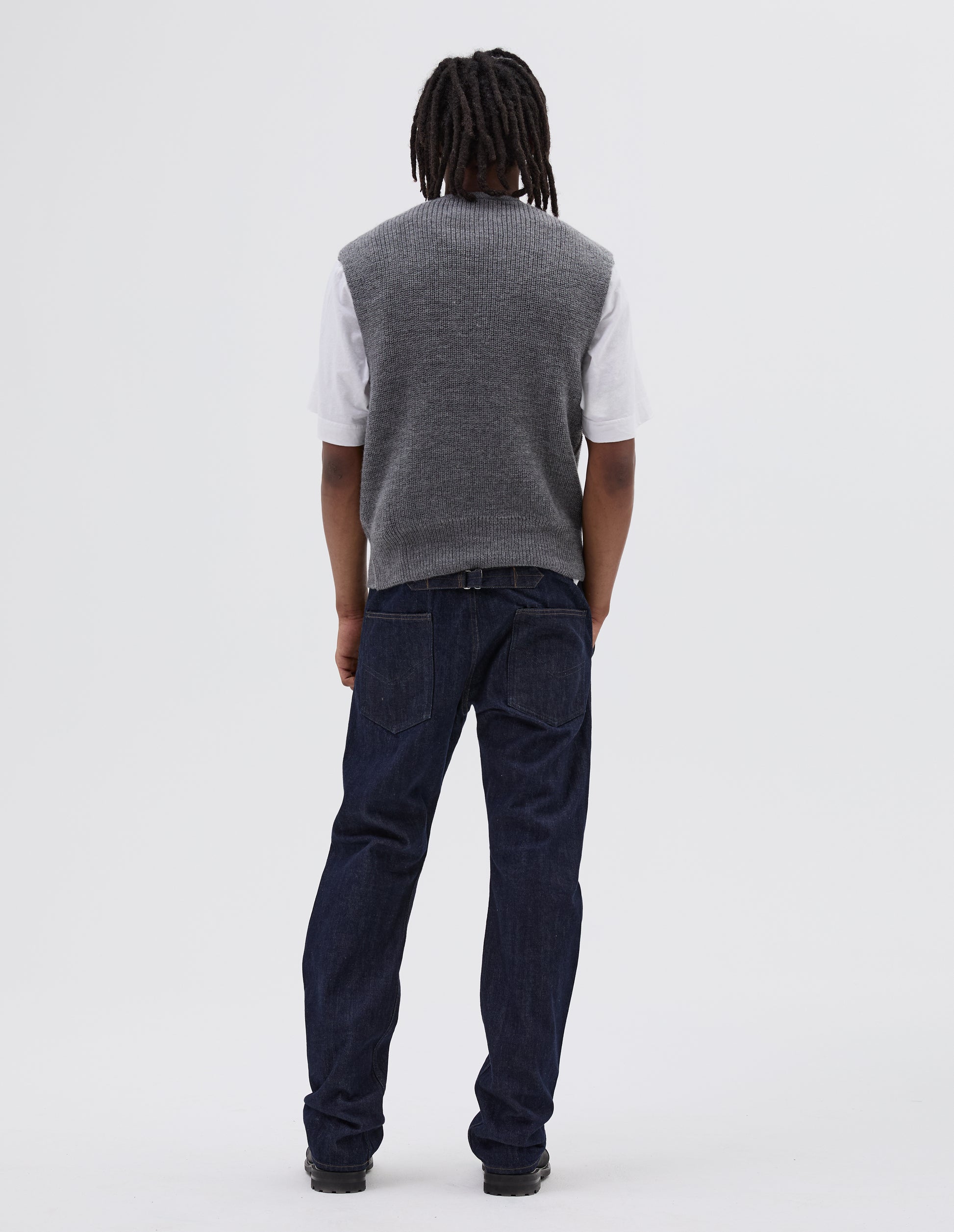 Full body image, back view of model wearing grey sleeveless ribbed v neck slipover jumper, with contrast rib on hem. Styled over white t shirt, with indigo denim jeans and black leather boots.