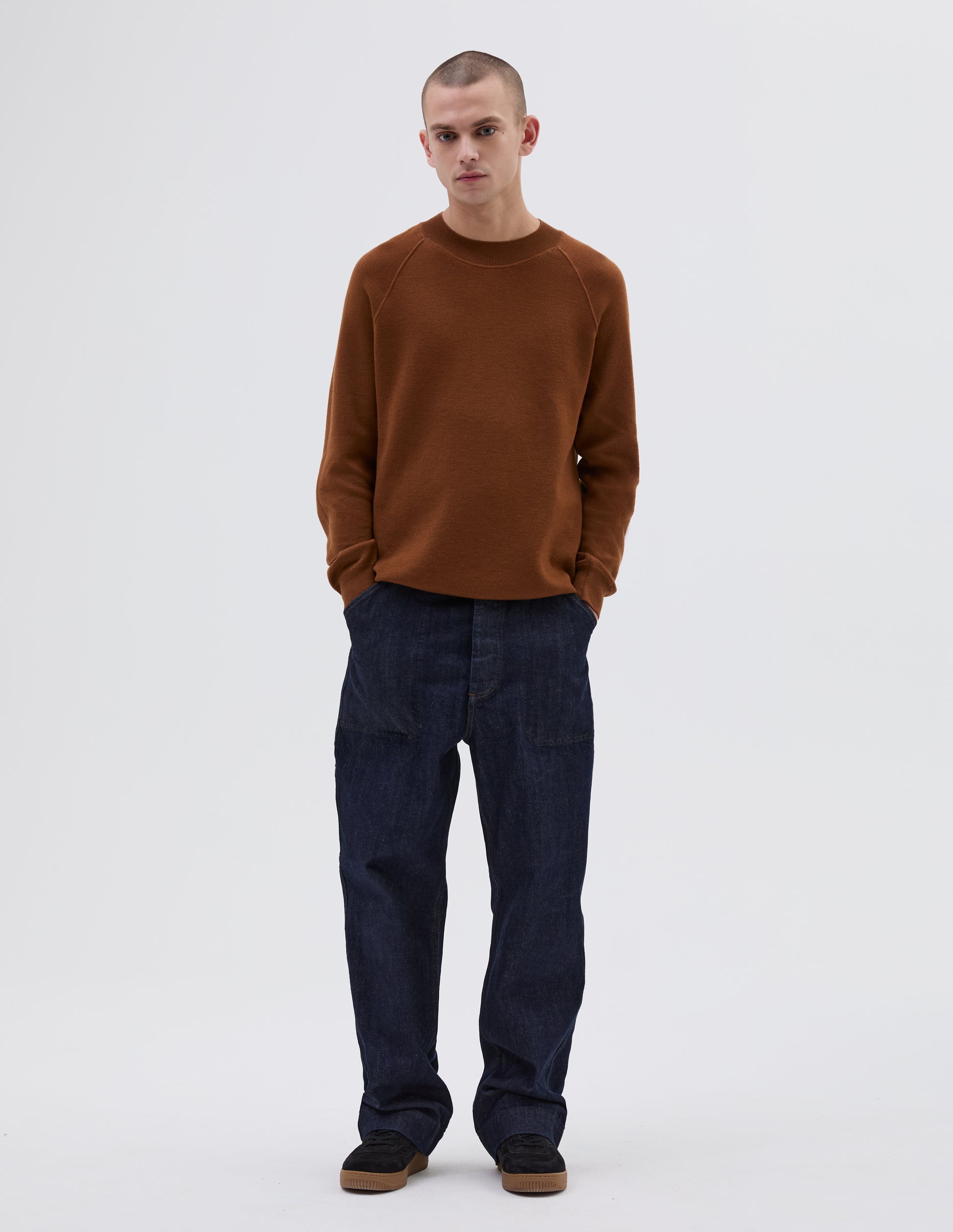 Full body image, front view of model wearing brown merino wool wide neck sweatshirt, with raglan sleeve, flatlock stitch detailing on seams and contrasting rib on neck, cuffs and hem. Styled with indigo blue jeans and black suede trainers.