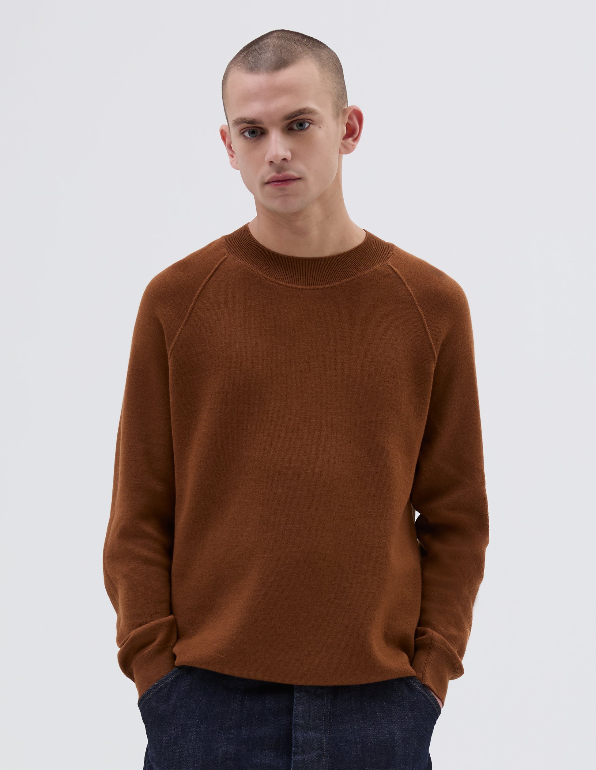 Cropped body image, front view of model wearing brown merino wool wide neck sweatshirt, with raglan sleeve, flatlock stitch detailing on seams and contrasting rib on neck, cuffs and hem. Styled with indigo blue jeans.