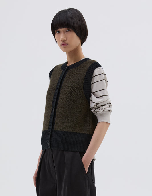 Cropped body image, side view of model wearing dark green and black contrast trim waistcoat, with contrast rib detailing and deep hem, finished with corozo brace button fastening. Styled over white and black striped long sleeve t shirt, with black pleated trousers.
